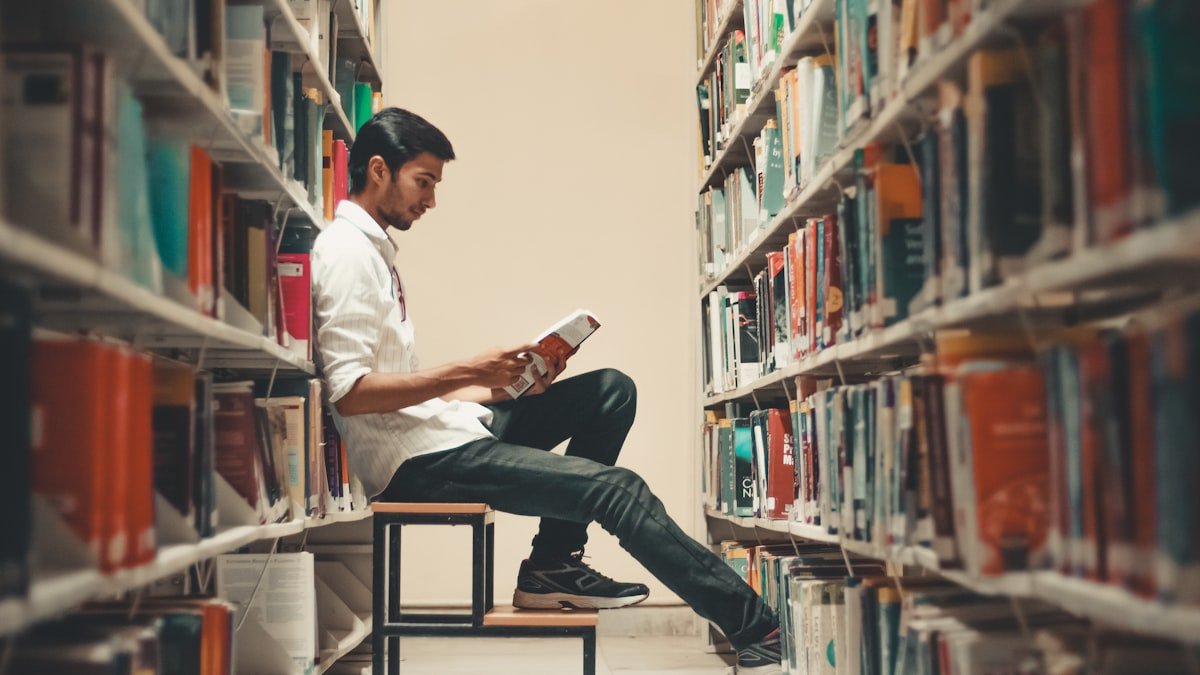 A man sitting and leaning near a bookshelf while reading a self-improvement book