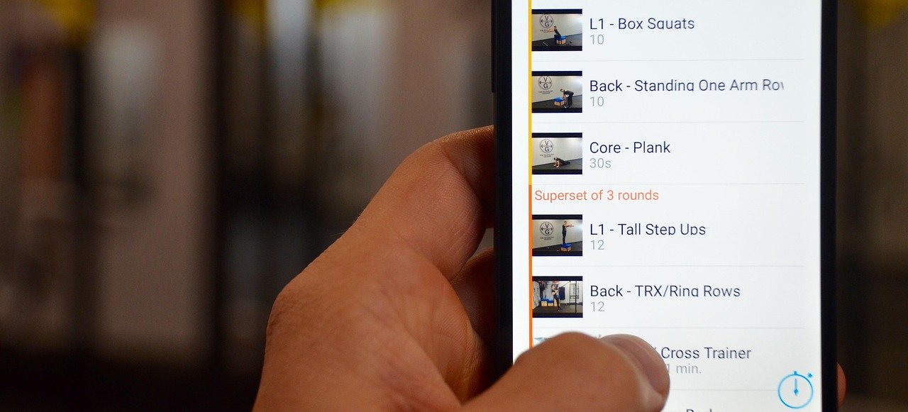 A hand holding a smartphone displaying a workout tracking app with an exercise list.