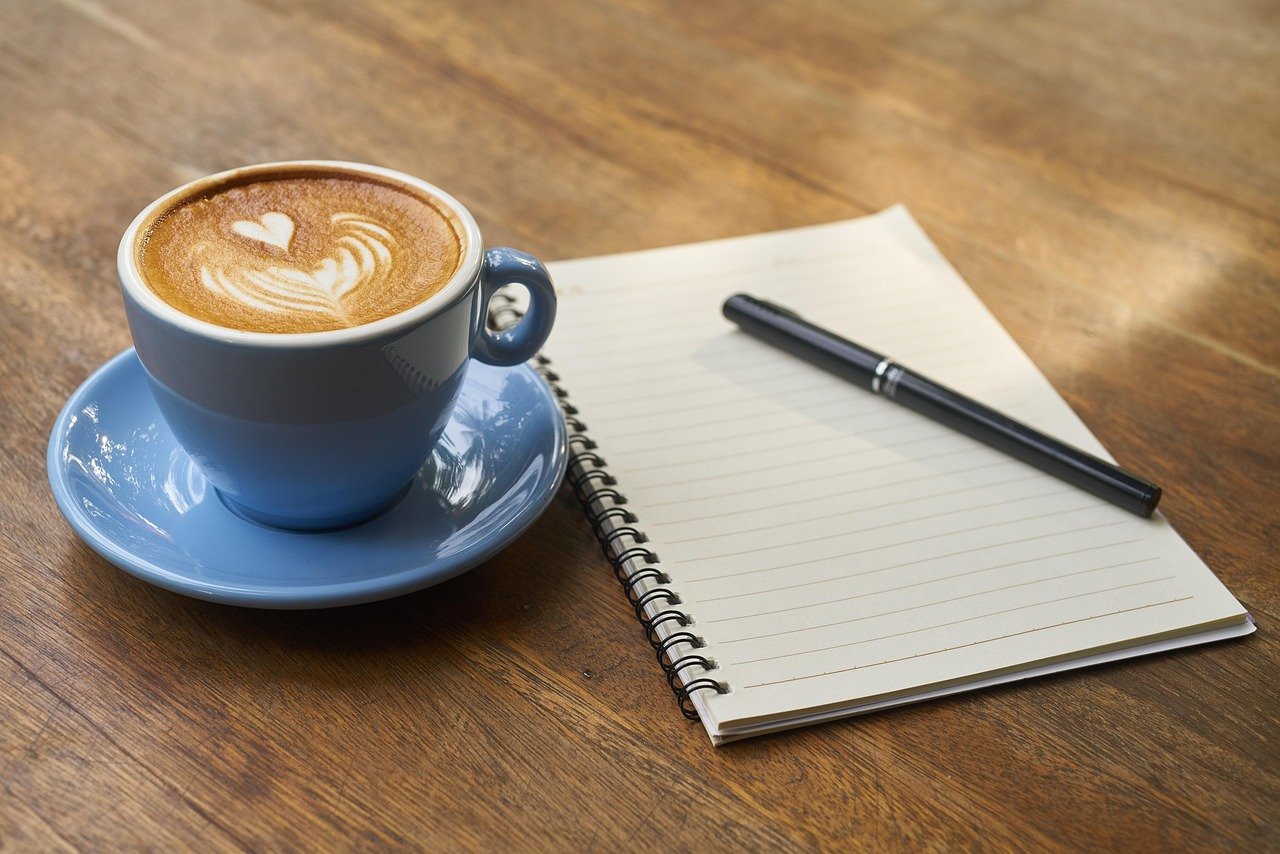 A cup of coffee next to an open notebook and pen, set up for a morning journaling session.