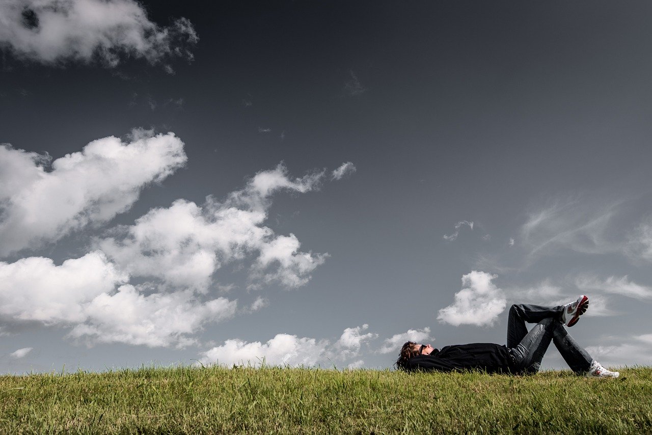 A man lying on his back on a grassy hill under a cloudy sky, resting