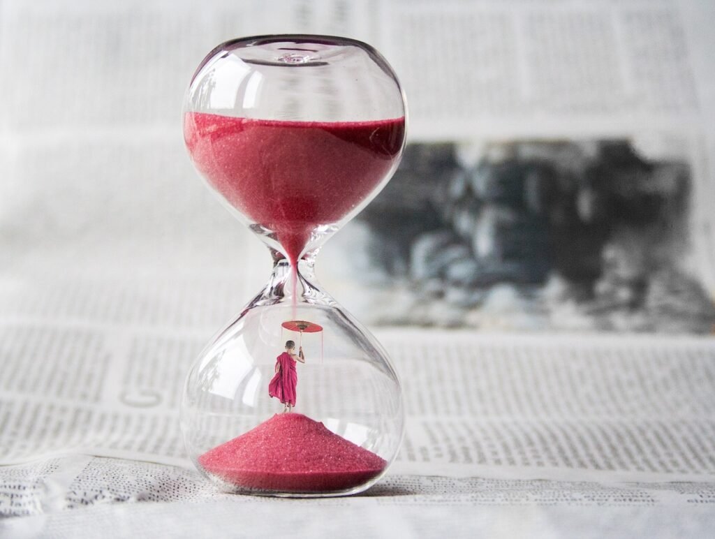An hourglass with pink sand running through it and a tiny robed figure standing inside, representing the question of how long to meditate